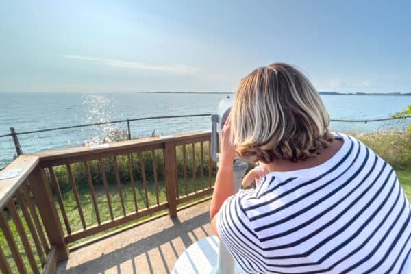 Woman looking through binoculars at Tibbetts Point Lighthouse with Lake Ontario in the background.